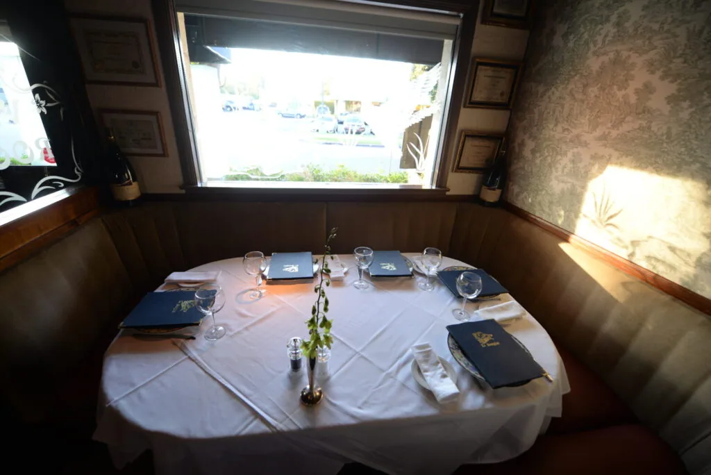 Fine dining table set for four with white tablecloth, blue napkins, glasses, and place settings in a wood-paneled booth with waterfront window view