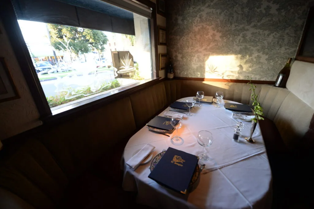Fine dining booth with white tablecloth, place settings, water glasses, and menu books, featuring a large window overlooking a waterfront promenade