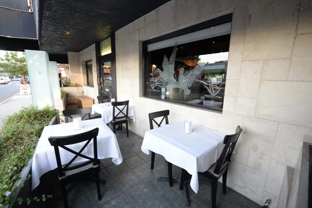 Outdoor patio dining area with white tablecloths, black chairs, and storefront windows