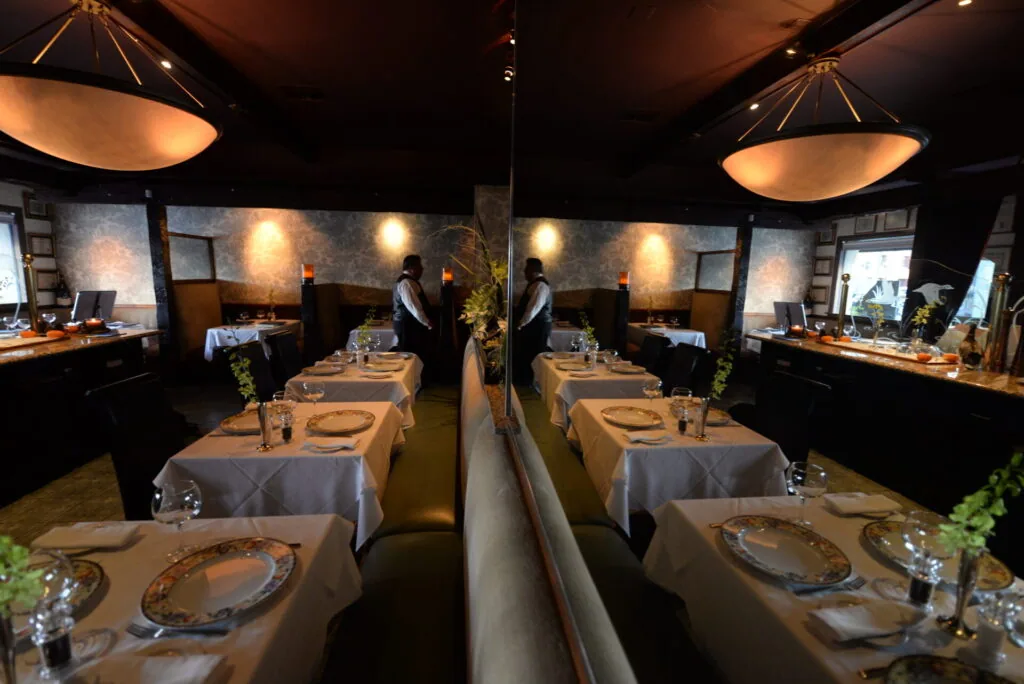 Upscale restaurant dining room with white tablecloth tables, elegant pendant lighting, and mirrored walls reflecting the sophisticated interior design.