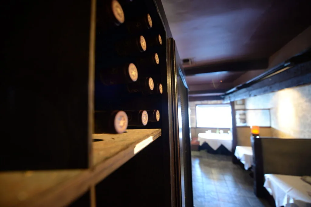 Interior of Le Sanglier restaurant with wooden beam in foreground and illuminated wall sconces, dining area visible in background