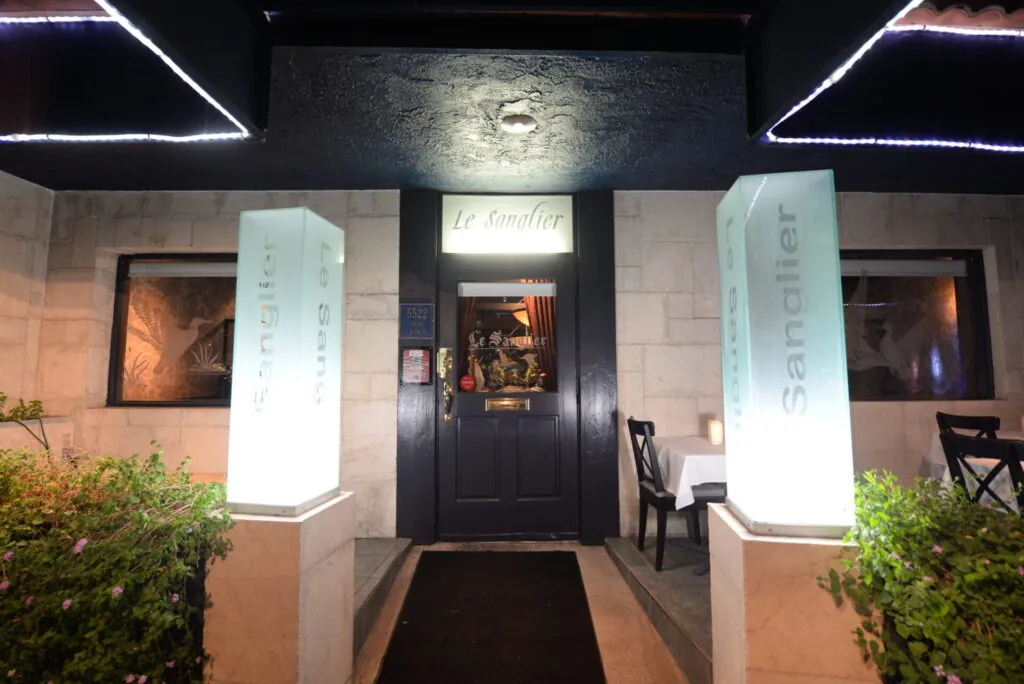 Restaurant entrance at night with illuminated pillars, neon ceiling lights, potted plants, and black door