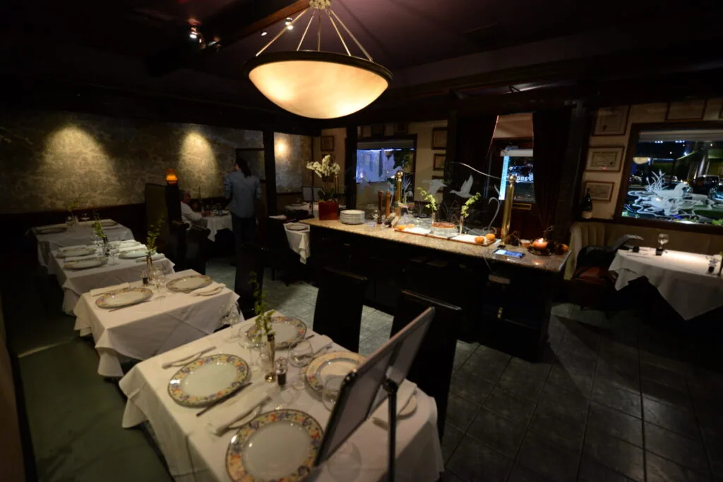 Upscale restaurant dining room with white tablecloth tables, place settings, pendant lighting, bar counter, and decorative elements