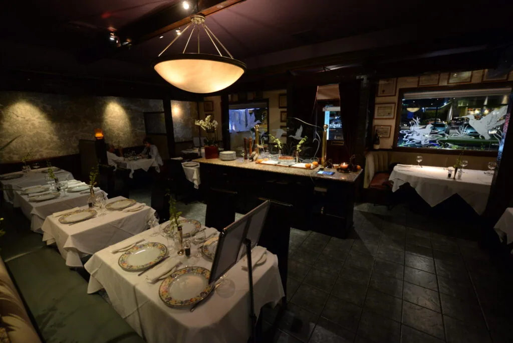 Upscale fine dining restaurant interior with white tablecloth tables, place settings with decorative plates, a brass pendant light fixture, bar area, and ambient lighting