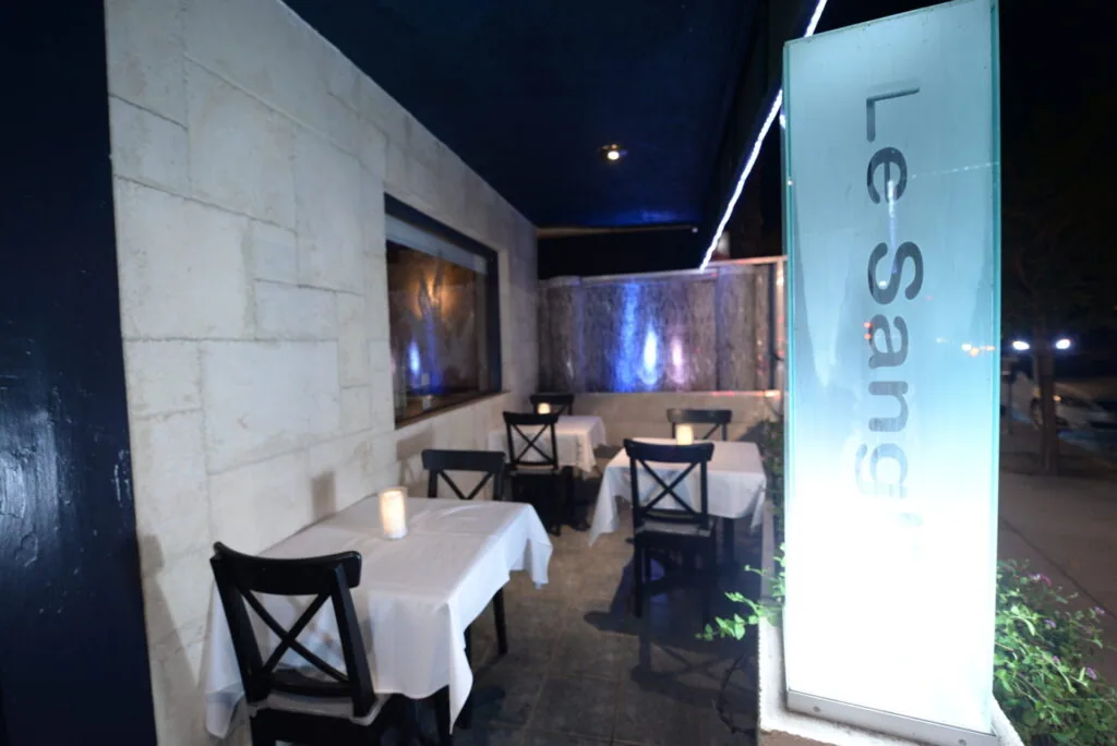 Nighttime exterior of Le Sanglier restaurant with illuminated signage, white tablecloths, and black chairs visible on the patio