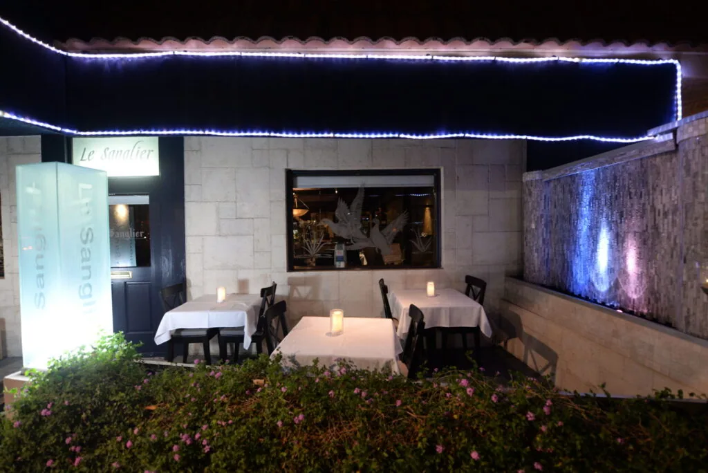 Outdoor restaurant seating at night with blue neon lighting, white tablecloths, black chairs, and flowering plants along the edge