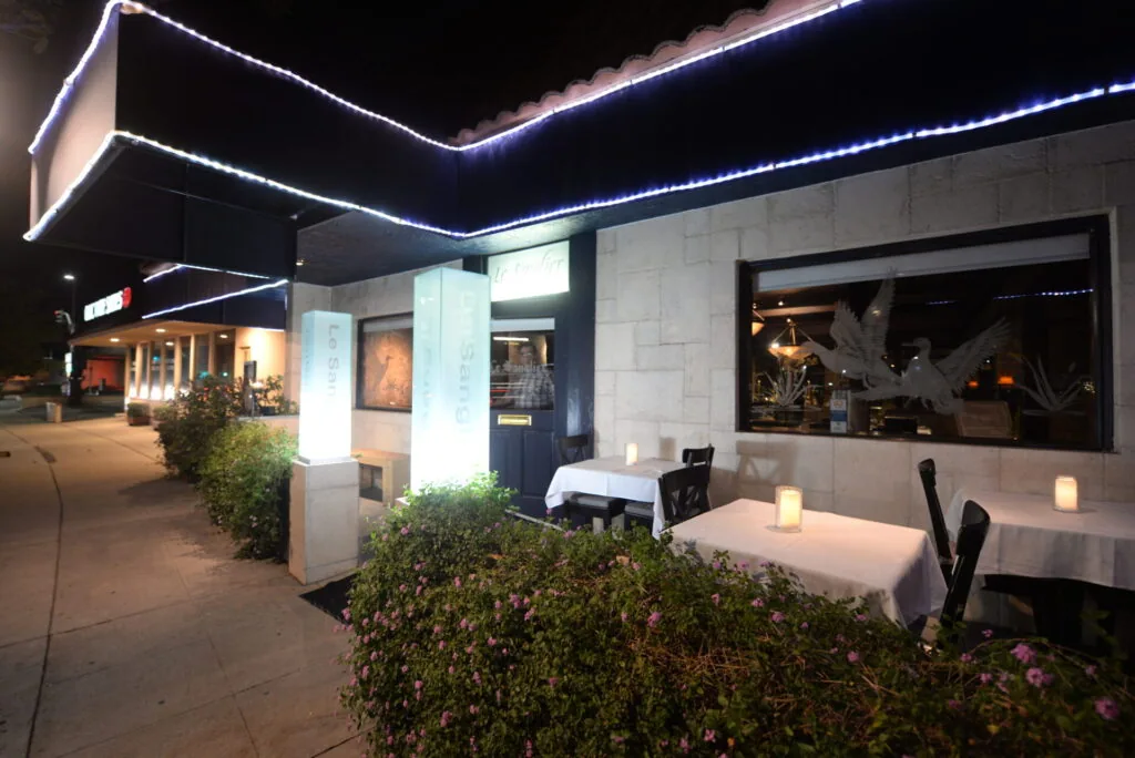 Modern restaurant exterior at night with illuminated ceiling edges, white tablecloths on outdoor seating area, potted plants, and large windows