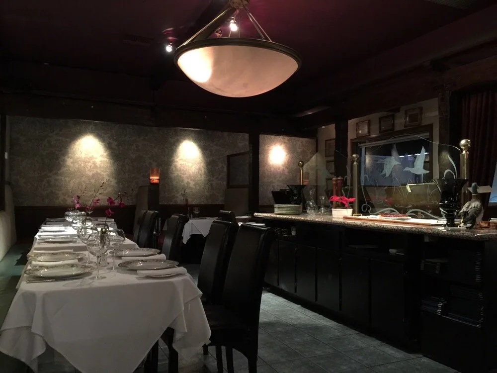 Fine dining room with white tablecloth-covered table, place settings, black chairs, pendant lighting, and bar counter with flowers and decorative items