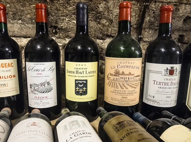 Row of wine bottles with red and black labels displayed against a stone wall, including Château selections and Bordeaux wines.