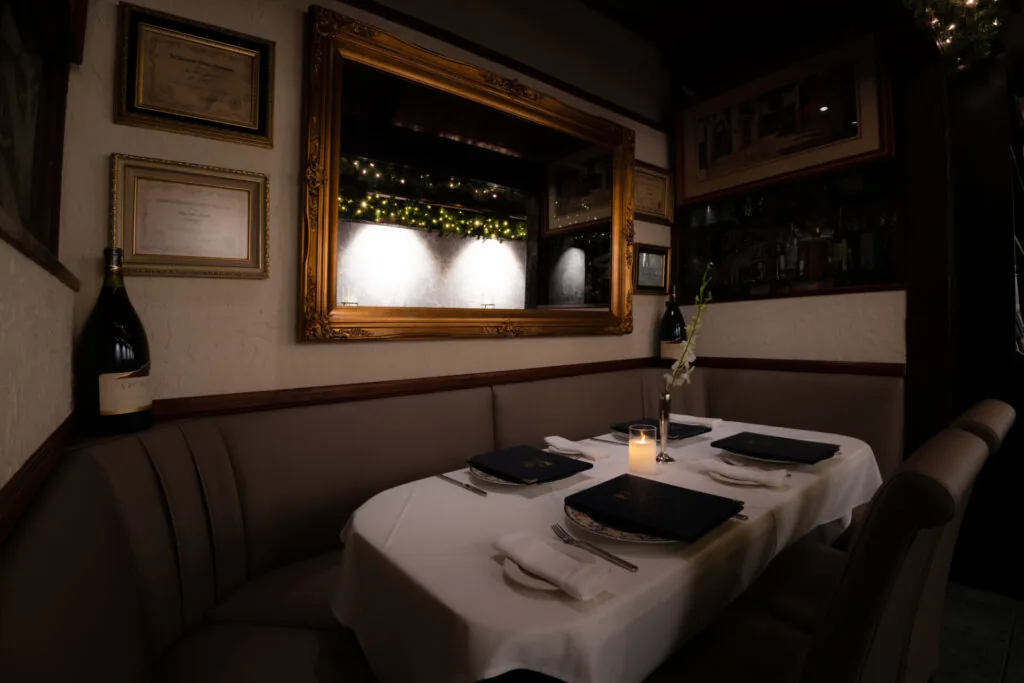 Intimate restaurant dining booth with white tablecloth, place settings, and wine bottle beside large ornate mirror with window view