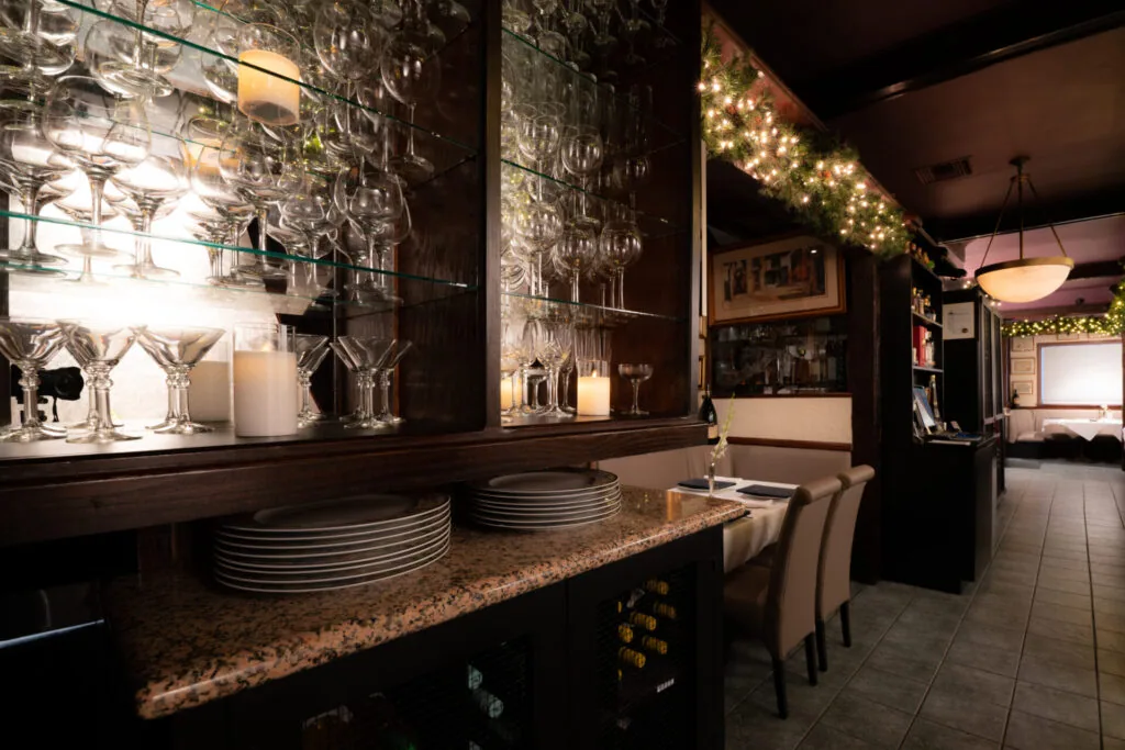 Restaurant bar counter with granite countertop, stacked plates, illuminated glassware display, and dining area beyond