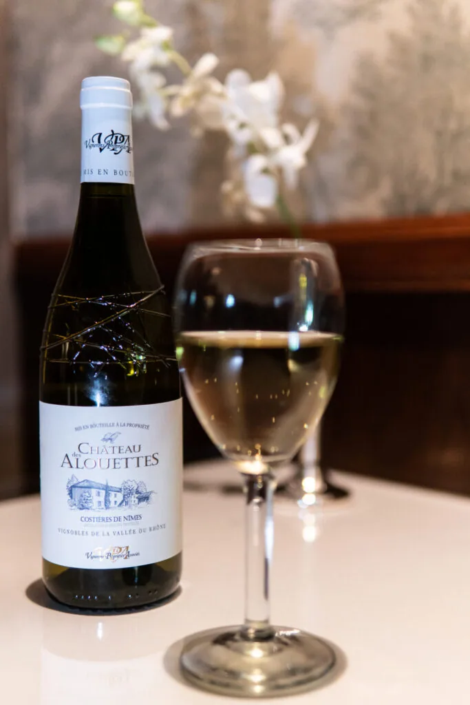 Wine bottle with white label next to a glass of pale wine on a table with blurred flowers in background