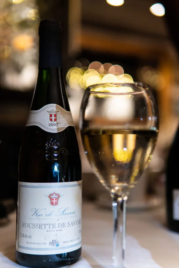 Wine bottle with white label next to a glass of white wine on table with warm blurred lights in background