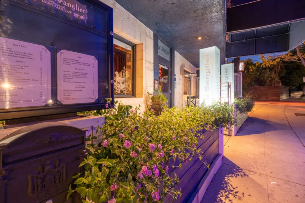 Restaurant storefront at dusk with illuminated menu boards and pink flowering plants in planters