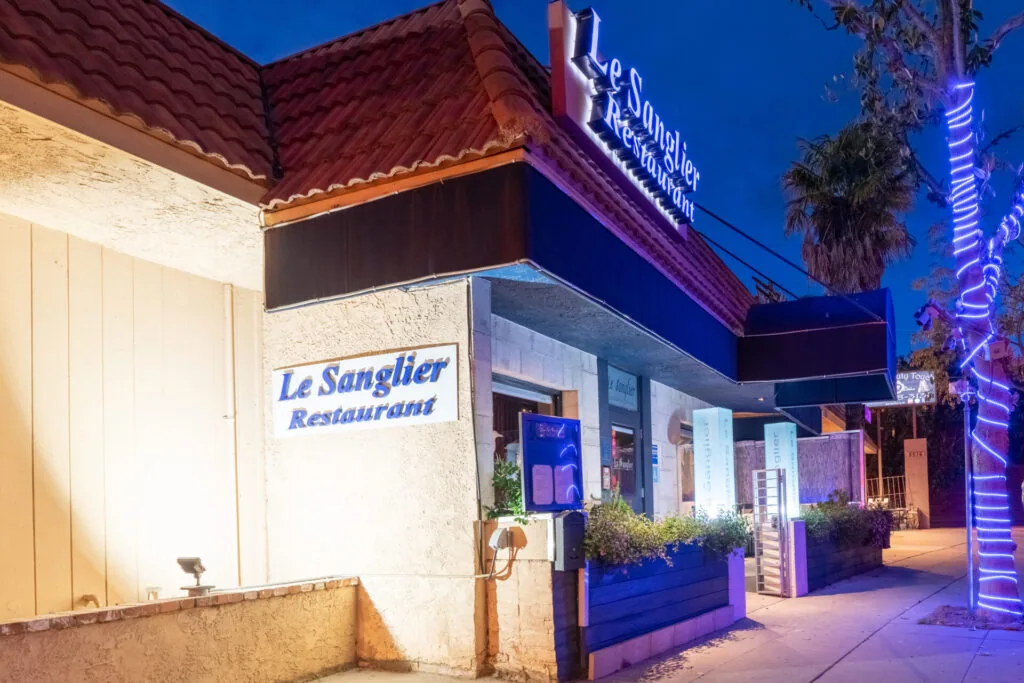 Restaurant storefront illuminated at night with neon signage and blue lighting, featuring white walls and landscaping.